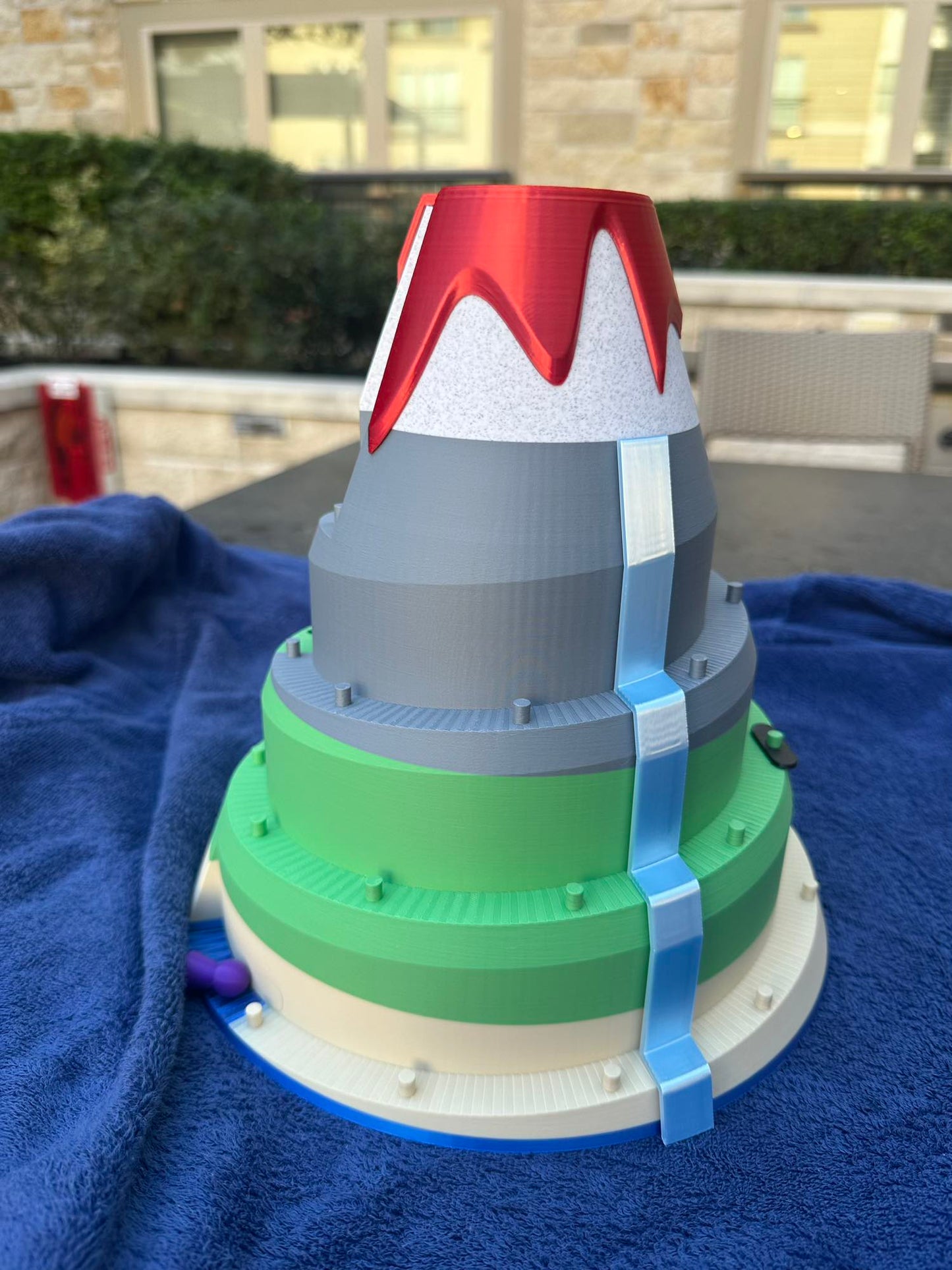 A 3D-printed volcano board game featuring layered tiers of green and gray slopes, a red lava peak, and a bright blue waterfall cascading down the mountain. Designed for racing pieces up the volcano in a fun, competitive tabletop game.