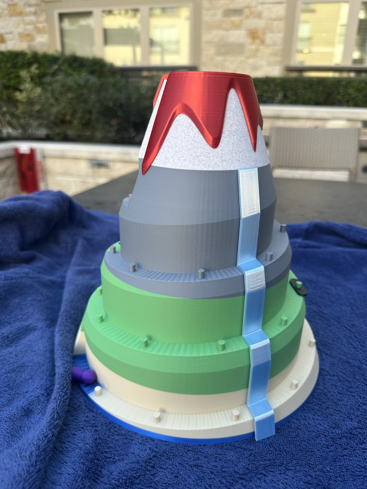 A 3D-printed volcano board game featuring layered tiers of green and gray slopes, a red lava peak, and a bright blue waterfall cascading down the mountain. Designed for racing pieces up the volcano in a fun, competitive tabletop game.