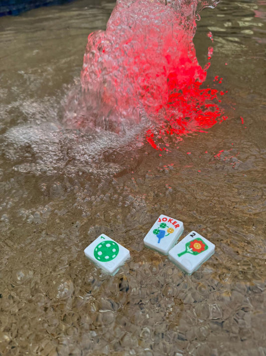 Pickleball Mahjong tiles floating in water beside a fountain with red light, demonstrating durable waterproof and pool-friendly design