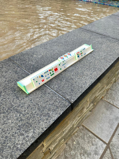 Green and white floating mahjong rack holding pickleball-themed mahjong tiles, displayed on a stone ledge next to a swimming pool