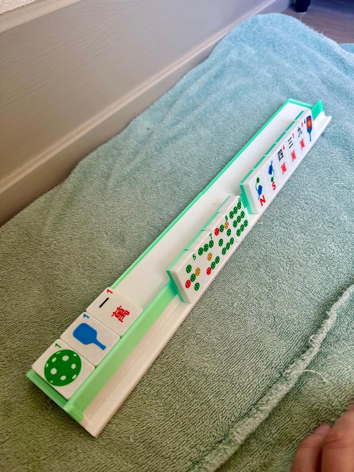 Close-up of green and white travel mahjong rack holding floating pickleball-themed mahjong tiles with colorful paddle, ball, and circle designs.