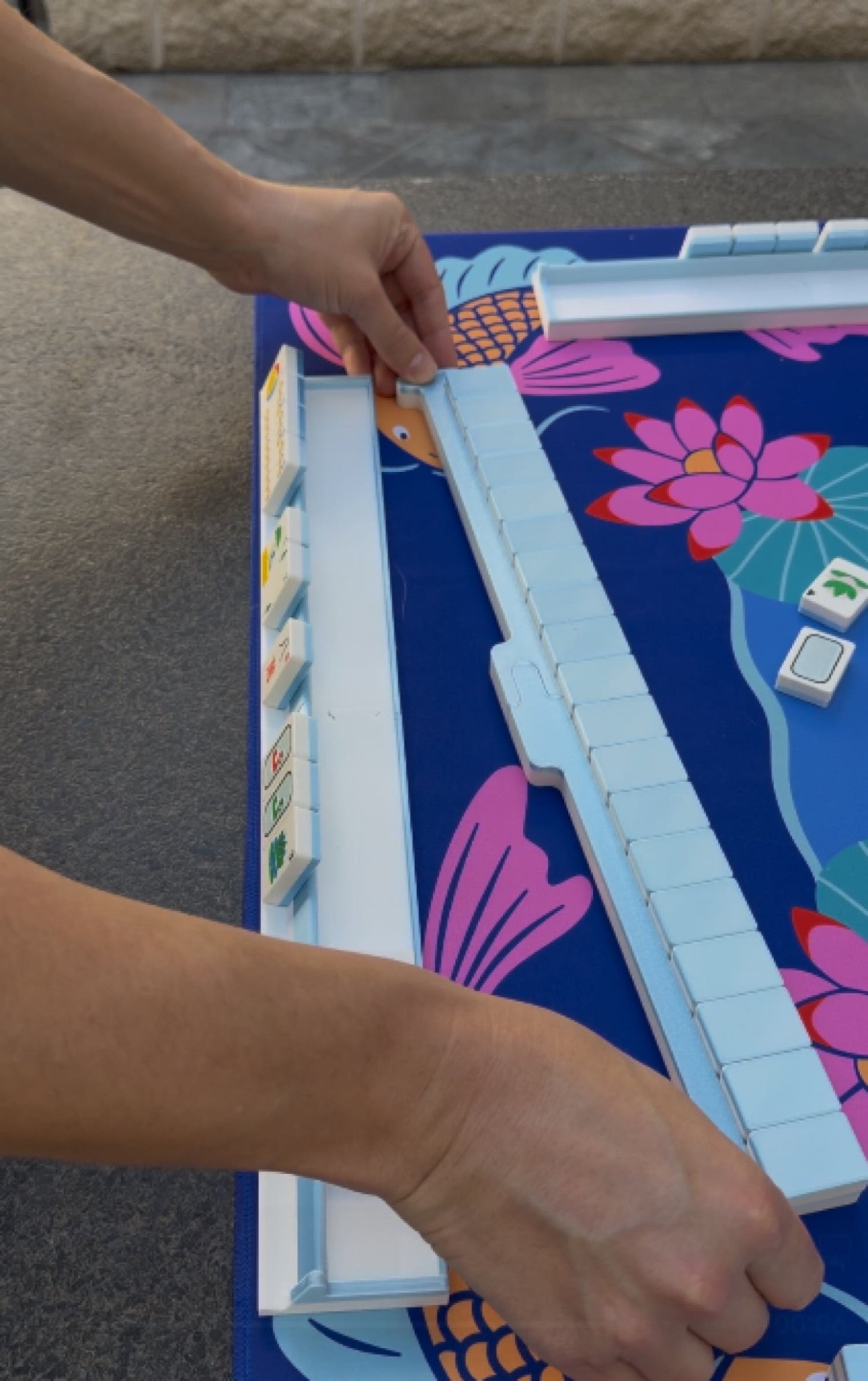 Hands assembling Spools pool mahjong racks on a poolside playing mat.
