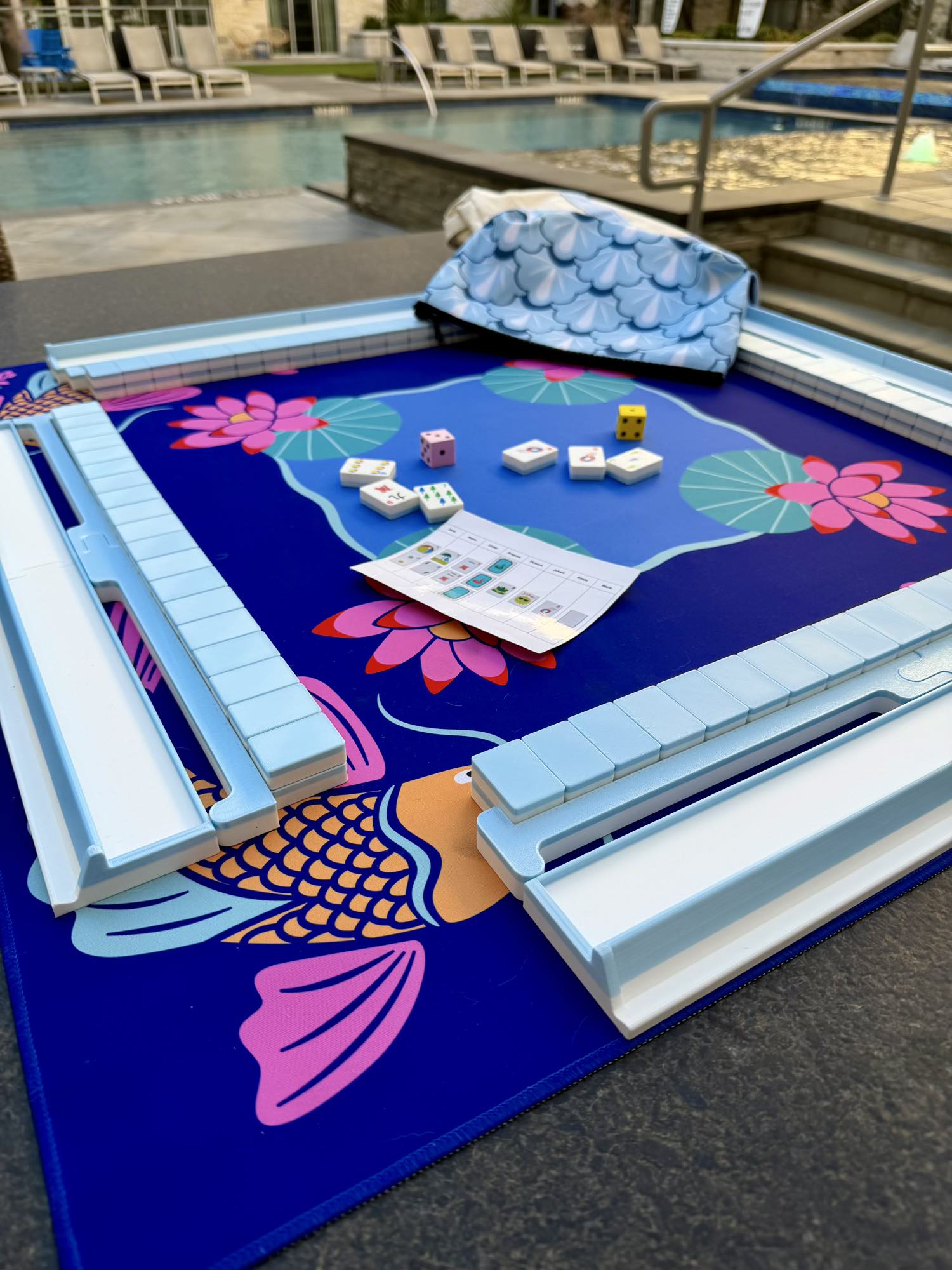Spools pool mahjong set with blue floating tiles and racks arranged poolside on a playing mat.
