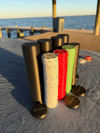 Spools travel poker chips and carrying cases arranged outdoors near the water, ready for play.

