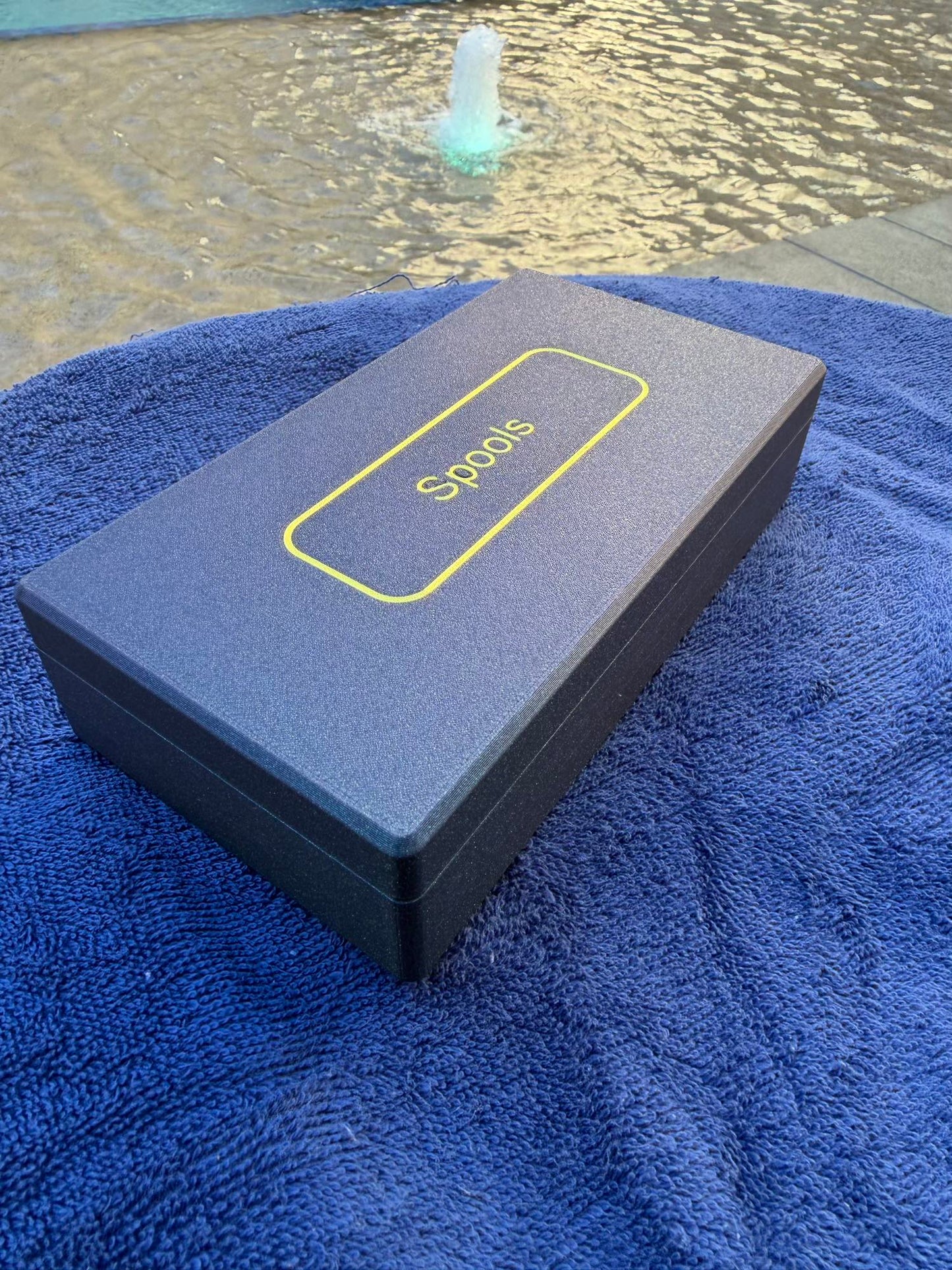Spools travel poker set closed in a slim gray case with logo, shown poolside on a blue towel.
