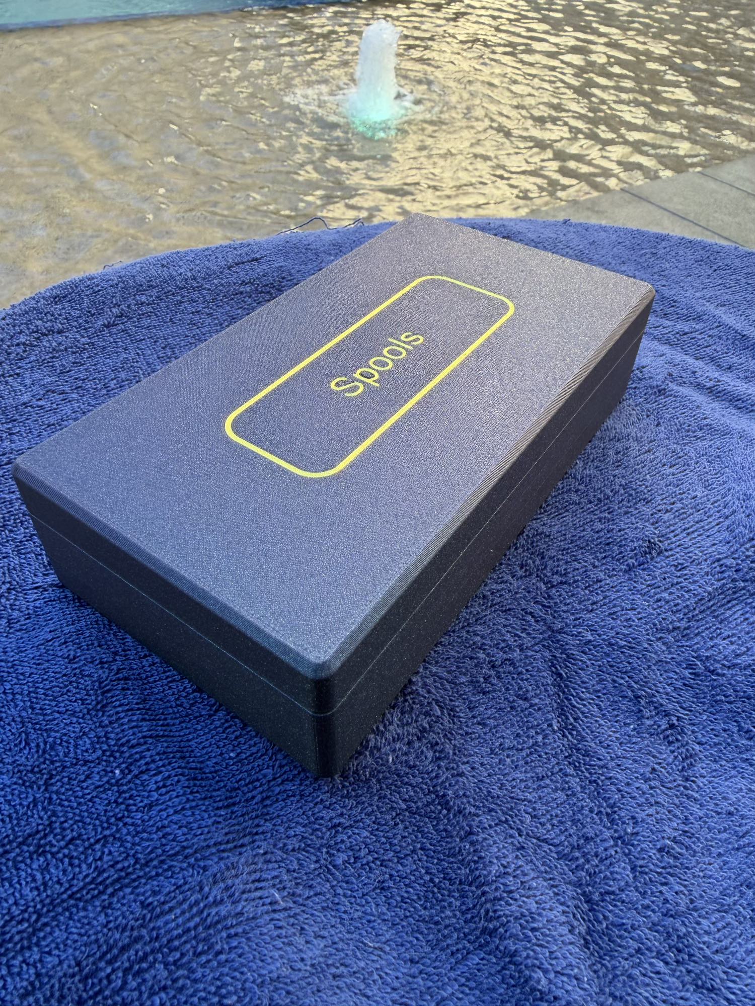 Spools travel poker set closed in a slim gray case with logo, shown poolside on a blue towel.

