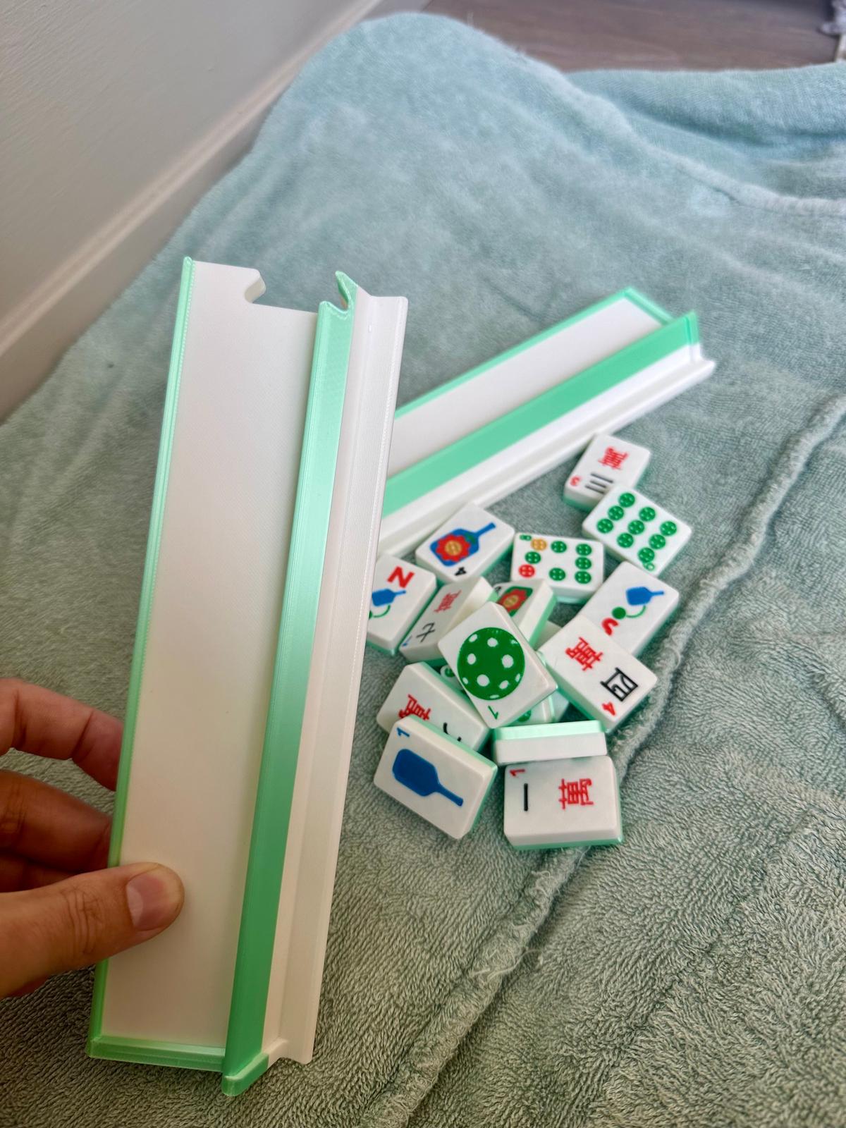 Hand holding mint green and white mahjong travel rack with floating pickleball-themed mahjong tiles scattered on a towel, including paddles, balls, and jokers
