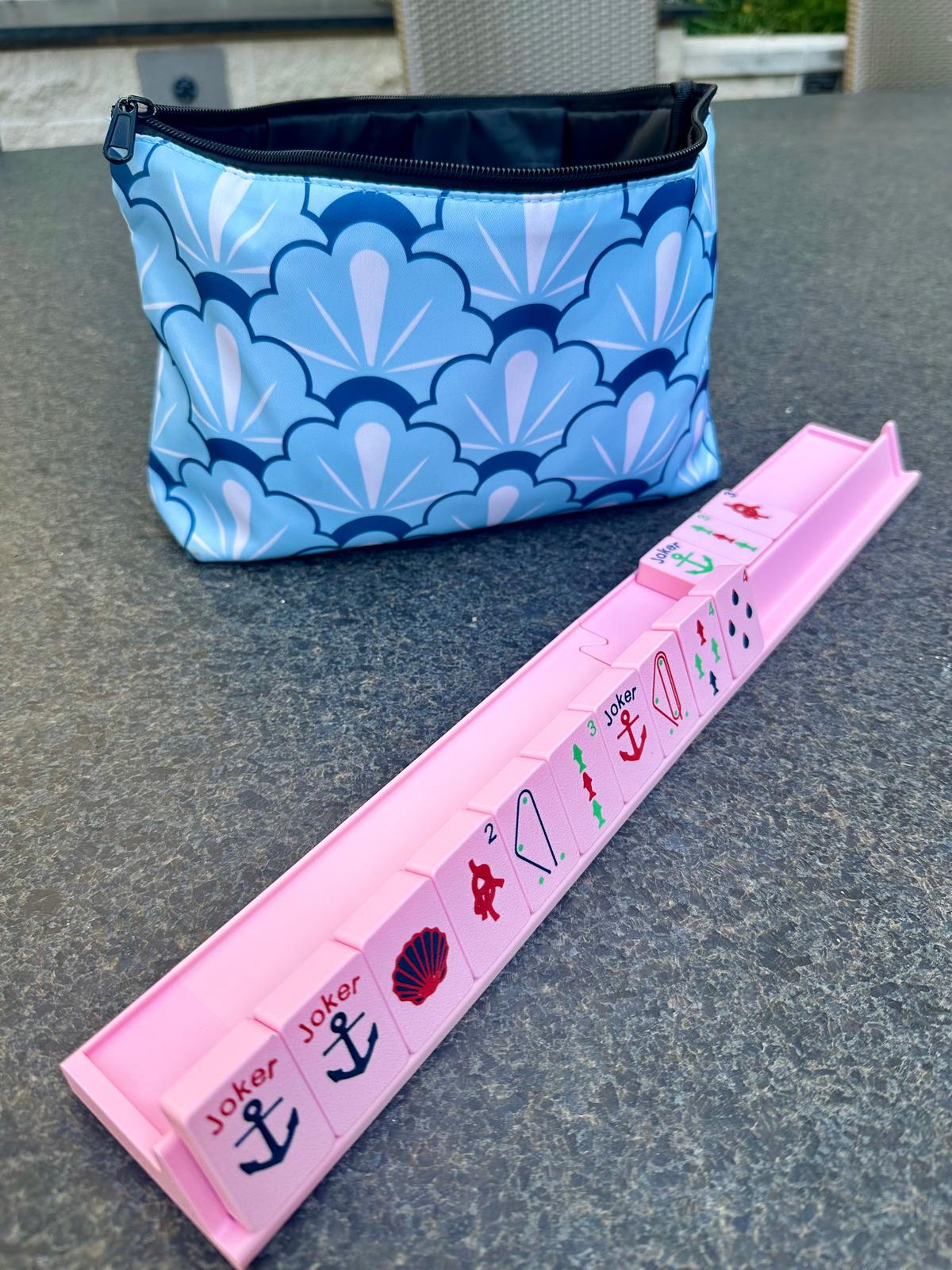 Pink floating mahjong travel rack holding nautical-themed tiles with anchor, seashell, and sailboat designs, shown next to a blue scallop-patterned carrying bag