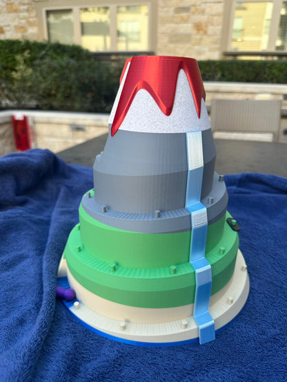 A 3D-printed volcano board game featuring layered tiers of green and gray slopes, a red lava peak, and a bright blue waterfall cascading down the mountain. Designed for racing pieces up the volcano in a fun, competitive tabletop game.